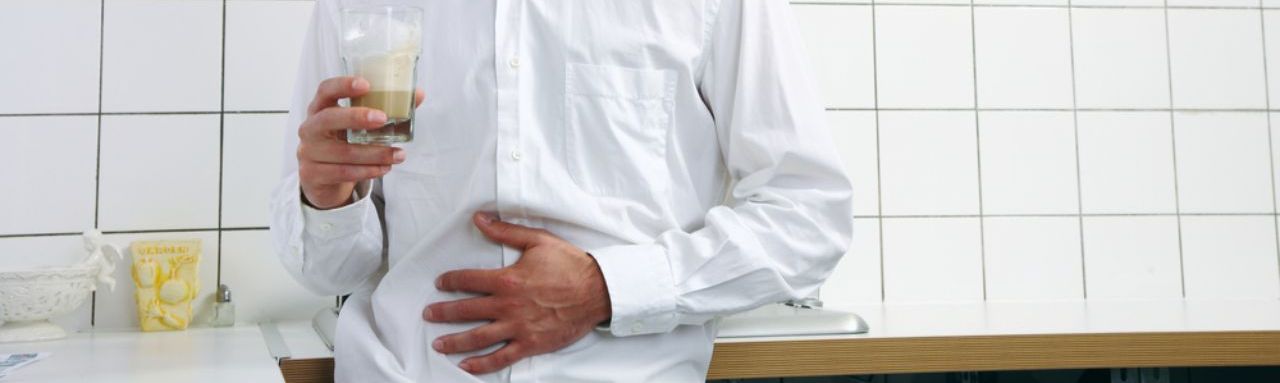 Un homme en chemise se tient dans une cuisine. Il se tiens le ventre mimant des douleurs à l'estomac