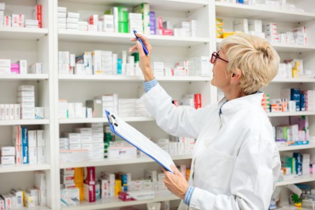 Une pharmacienne tenant un bloc note se tiens face à des étagères remplies de médicament
