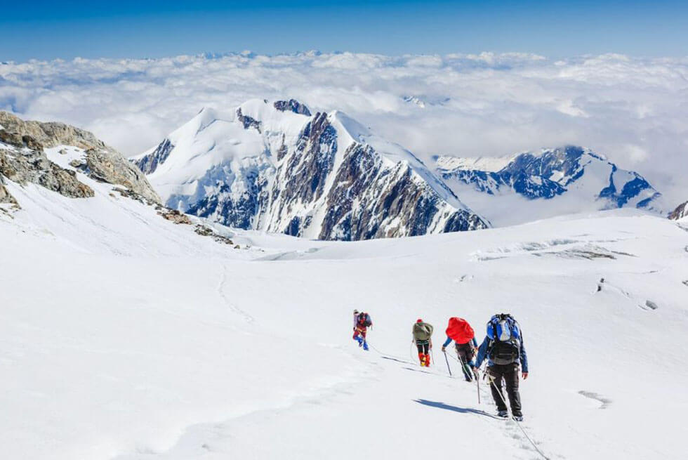 Une équipe de 4 personnes marchent à travers une plaine enneigée face à une montagne