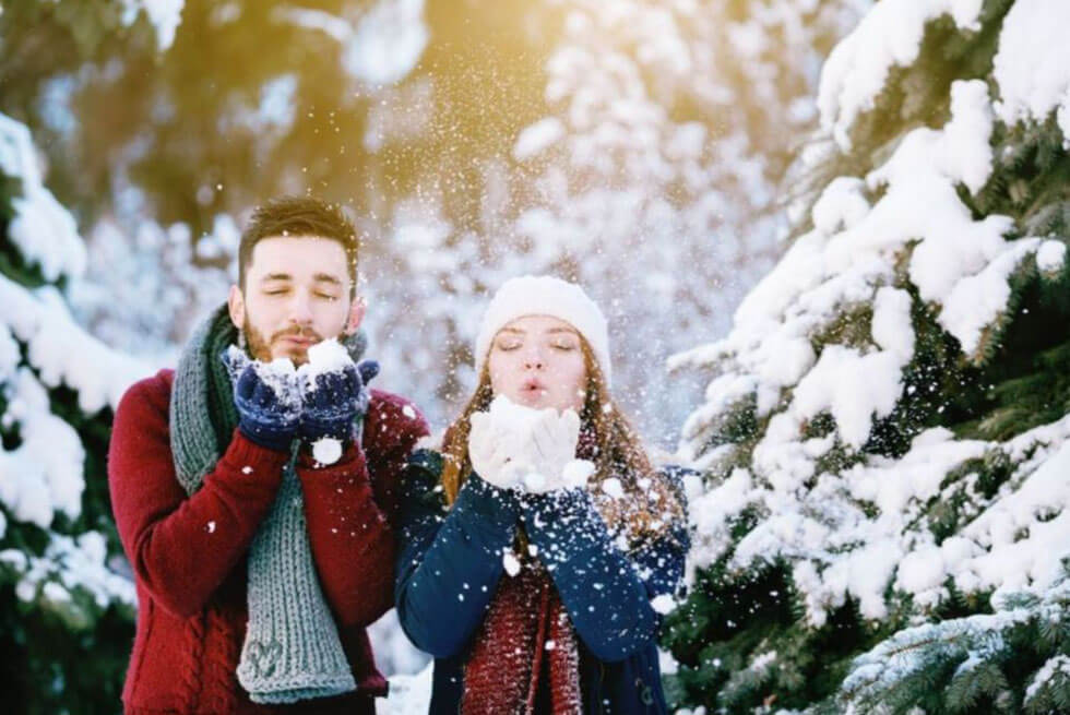 Un couple se tient dans une forêt enneigée et souffle sur un petit tas de neige dans leurs mains