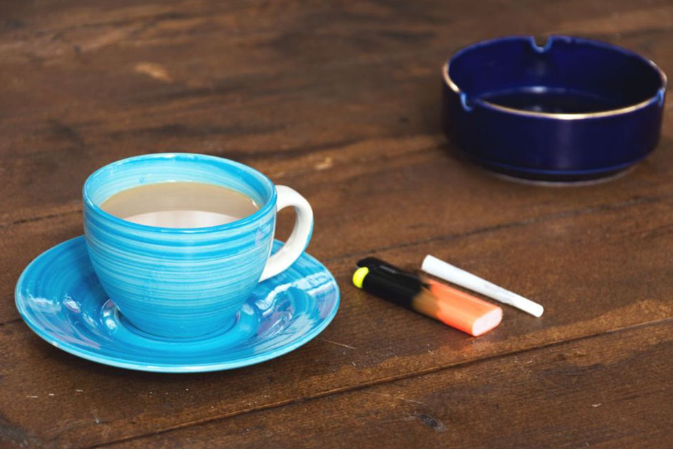 une tasse de café au lait, un briquet, une cigarette et un cendrier sont posé sur une table en bois