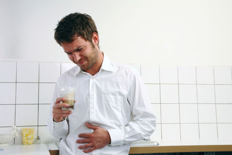 Un homme en chemise se tient dans une cuisine. Il se tiens le ventre mimant des douleurs à l'estomac