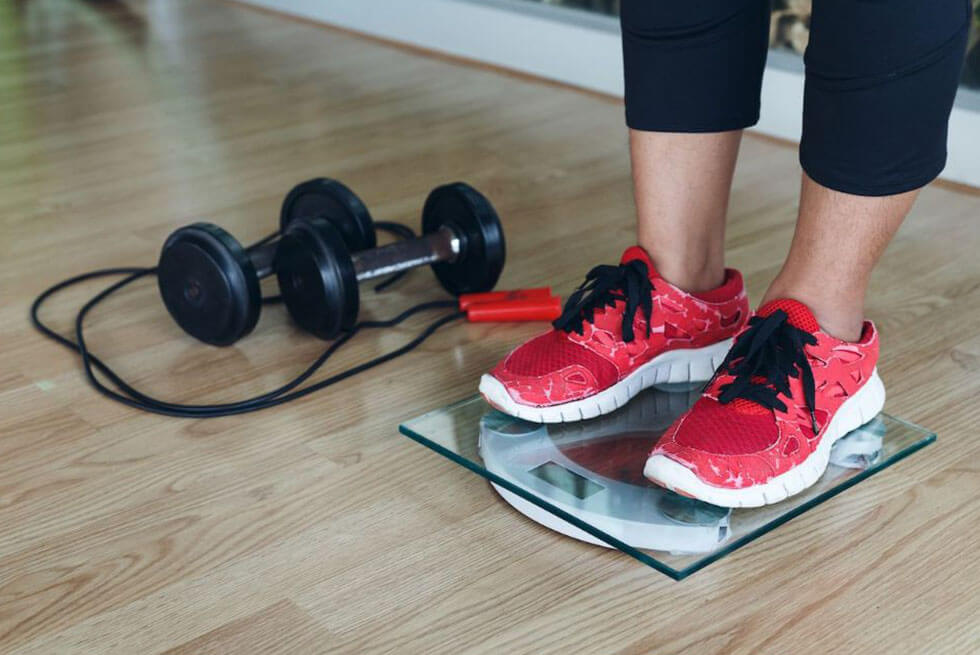 Une femme se pèse avec ses chaussures de sport, à coté d'elle est posé du matériel de fitness